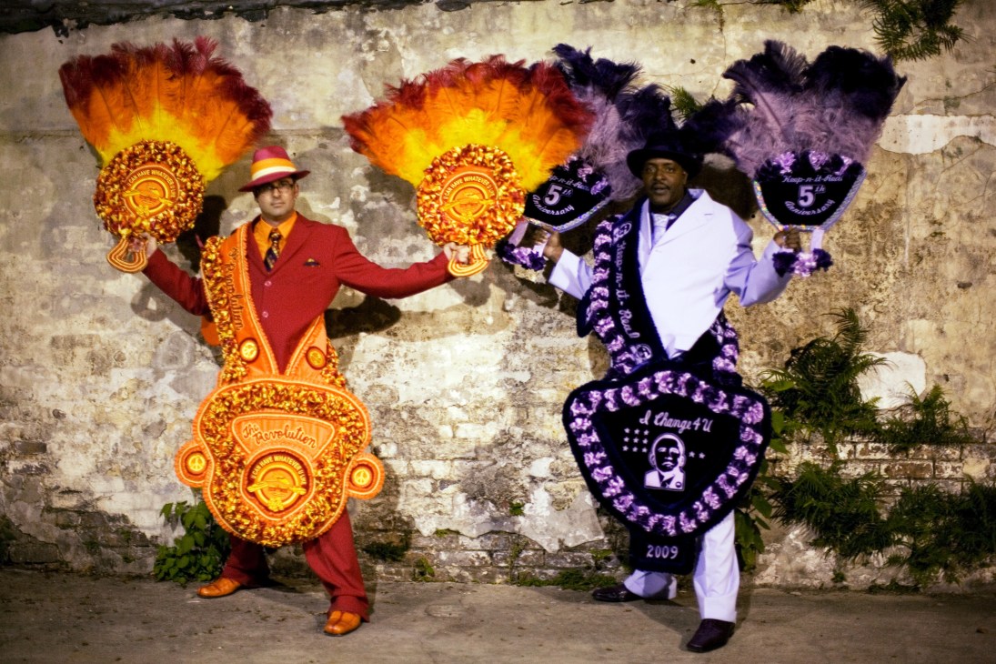 Two men in colorful suits and feathered headdresses perform traditional New Orleans second line dance against a weathered brick wall.