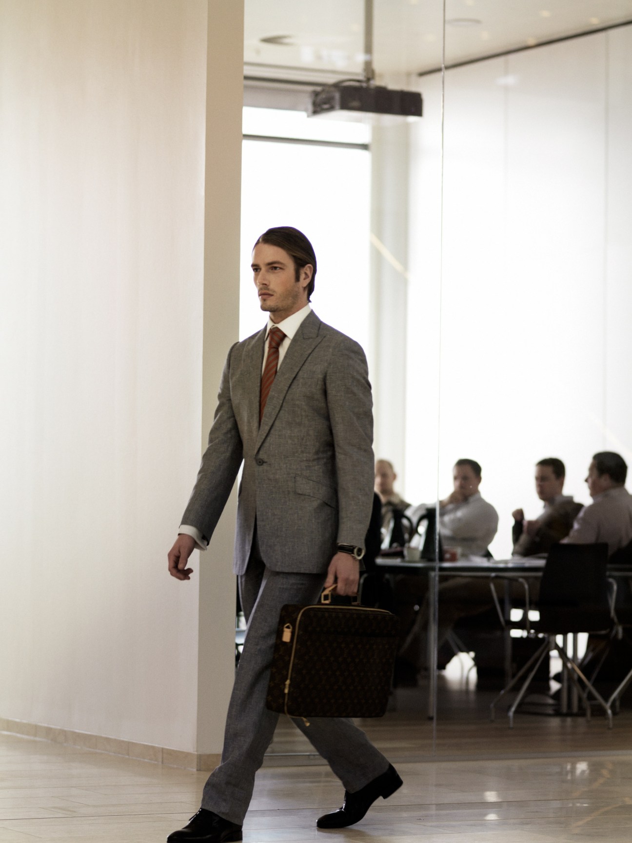 Man in grey wool suit walking through modern office corridor with briefcase.