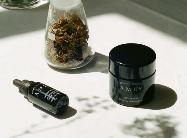 Amly skincare products displayed with dried botanicals in a glass jar.