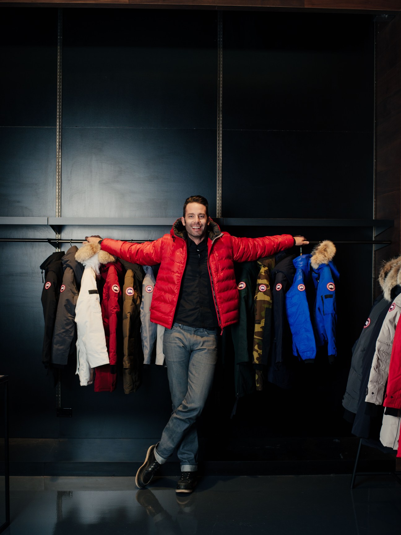 Man in red Canada Goose parka with arms spread wide among colorful coats on display rack