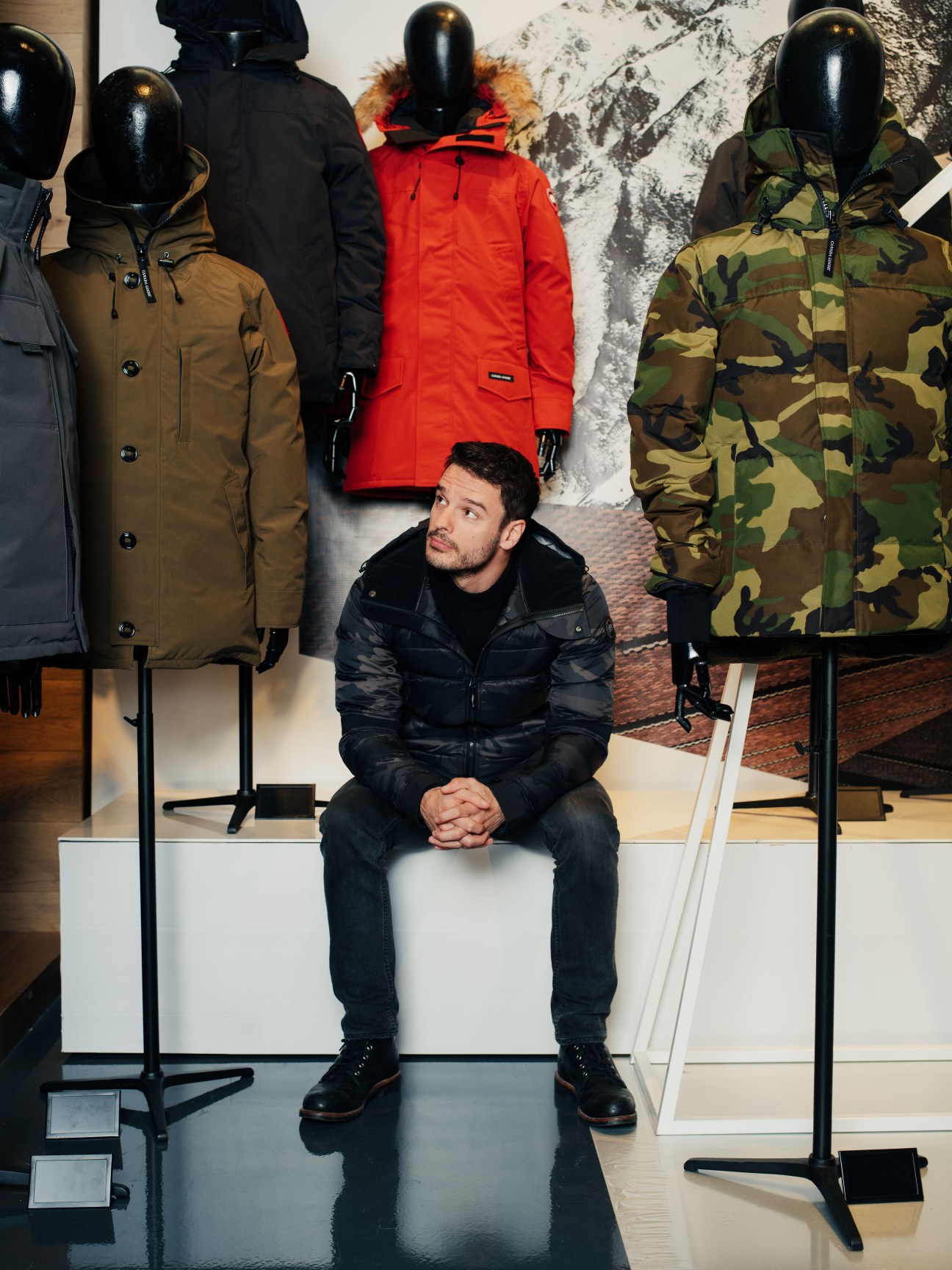Man sitting on white bench surrounded by Canada Goose parkas on mannequins in various colors including red, black, tan, and camouflage