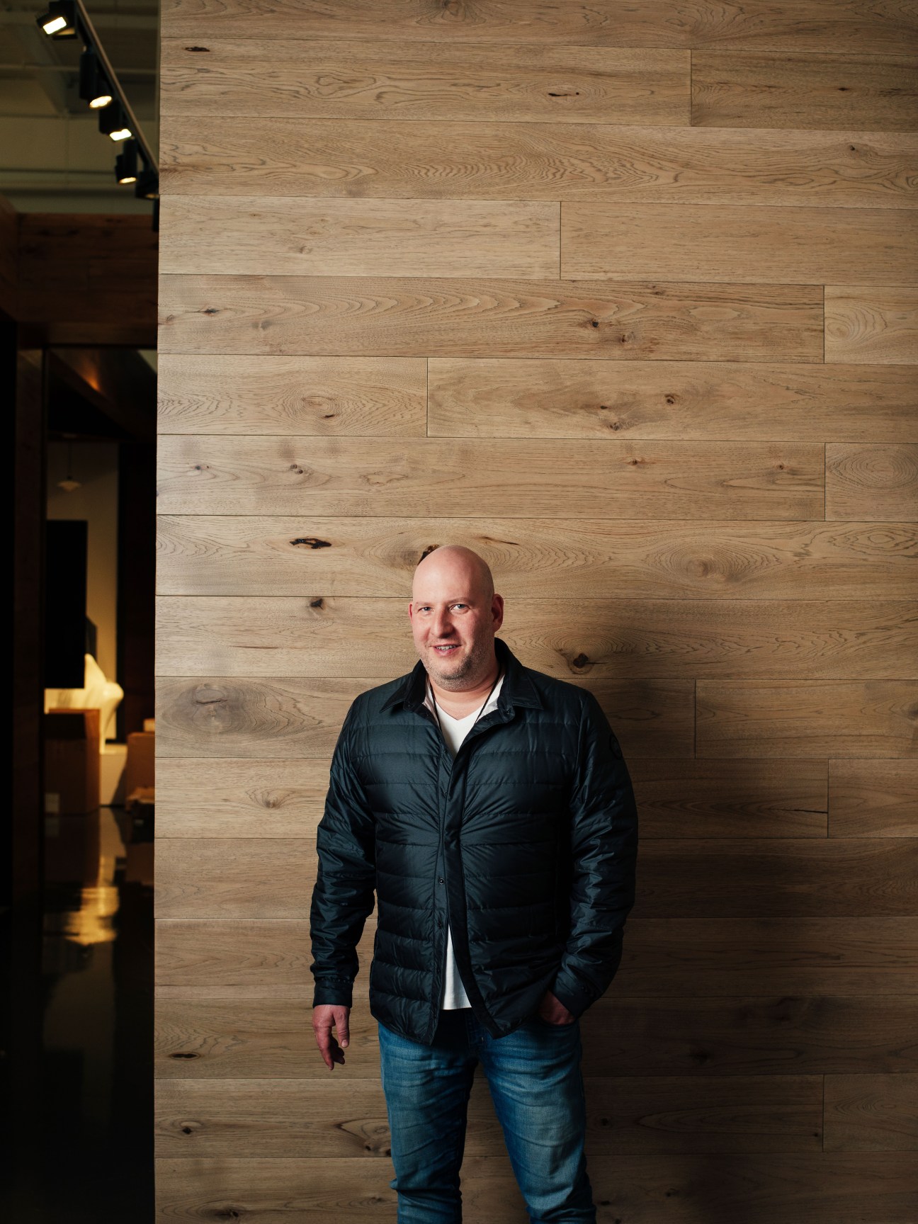 Dani Reiss, president and CEO of Canada Goose, wearing a black down jacket stands against a wooden wall at company headquarters.