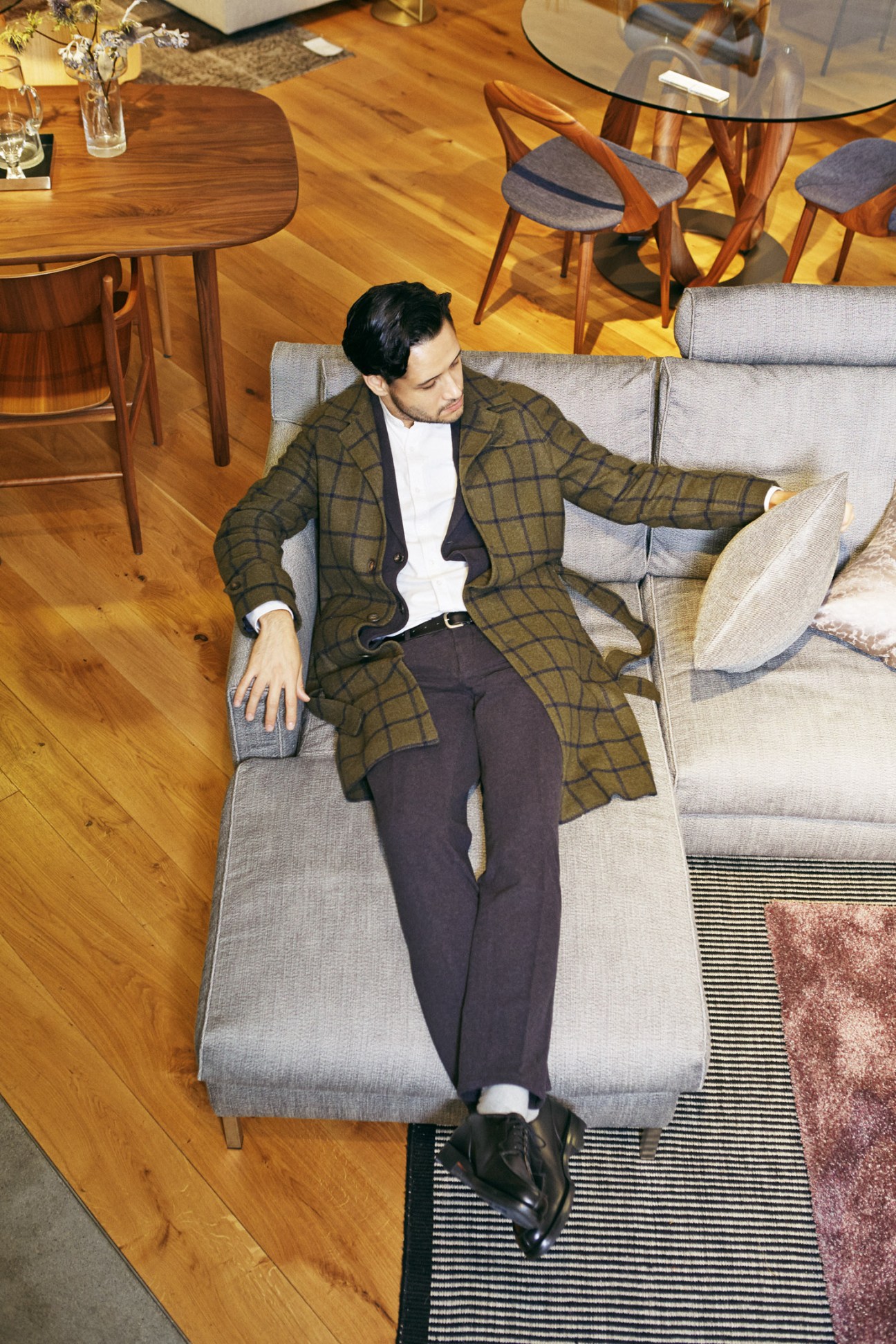 Man in plaid coat and dark trousers reclining on gray sofa in modern interior with wooden dining table and chairs.