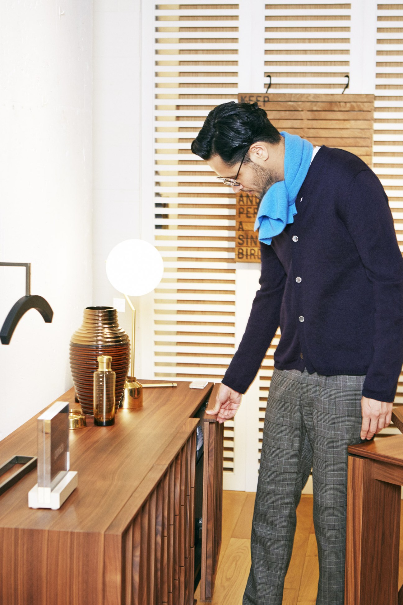 Man in navy cardigan and grey trousers leaning over wooden cabinet in minimalist interior