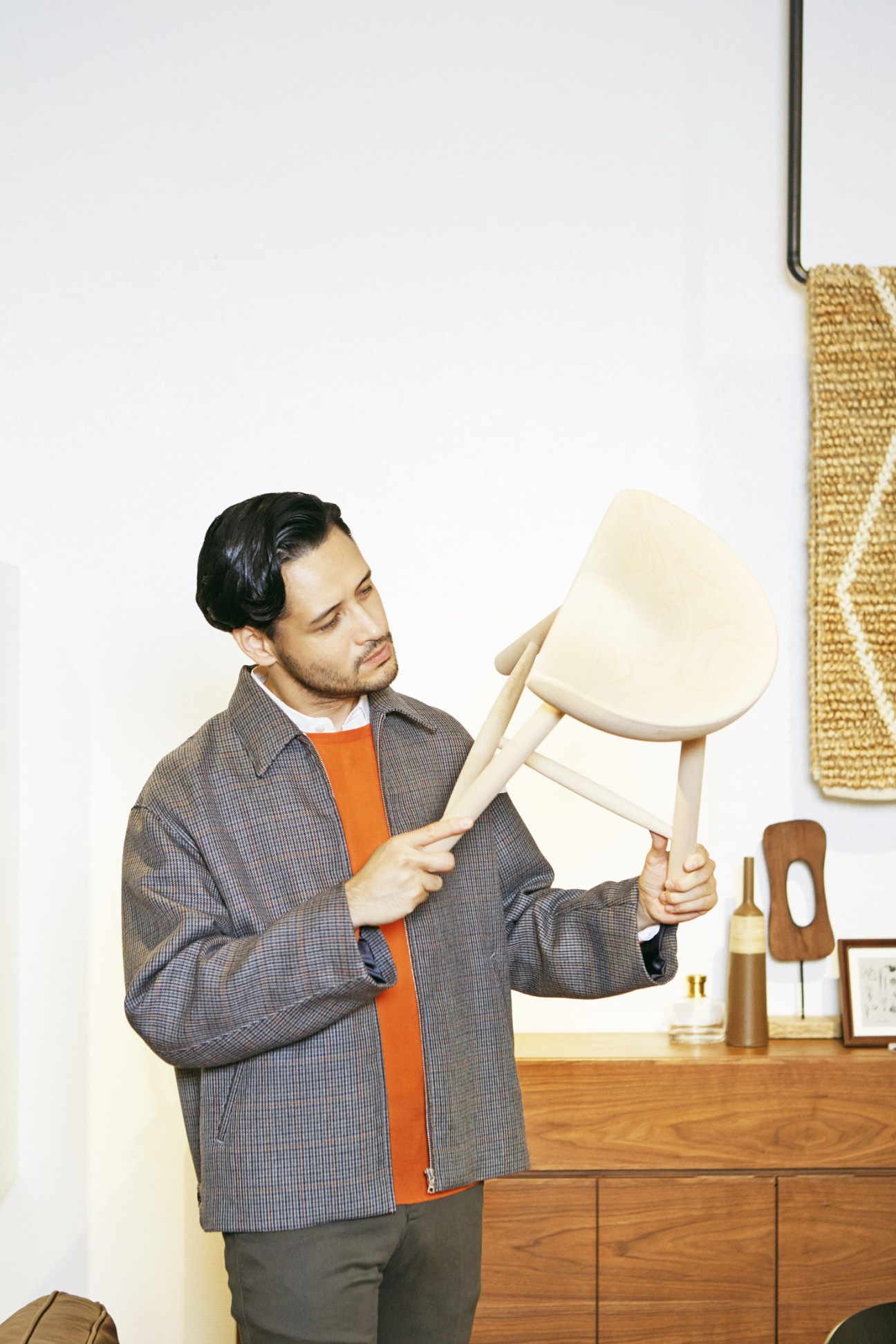 Man in Auralee jacket and John Smedley jumper examining a cream-colored chair in a minimalist interior.
