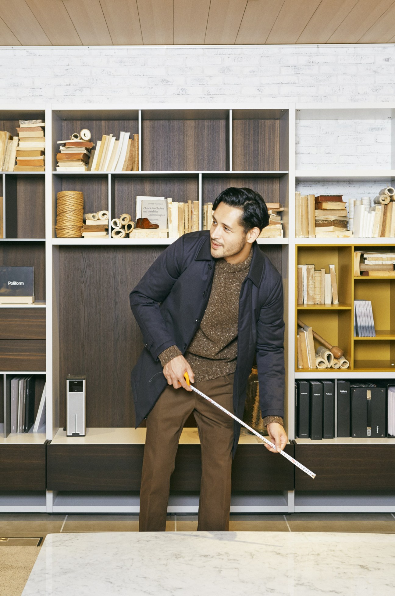 Man in navy Valstar coat and Finisterre jumper holding tape measure in front of Poliform shelving system