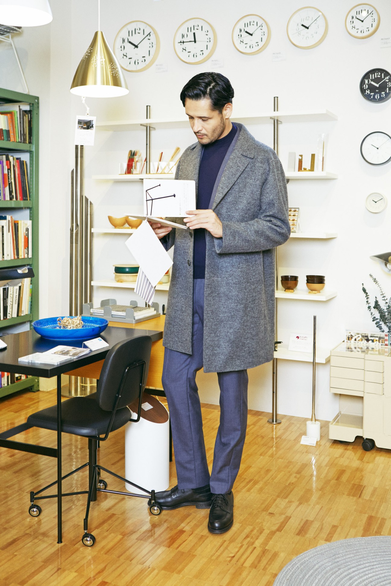Man in gray coat and navy Prada suit reviewing documents at home office desk with shelving and clocks.
