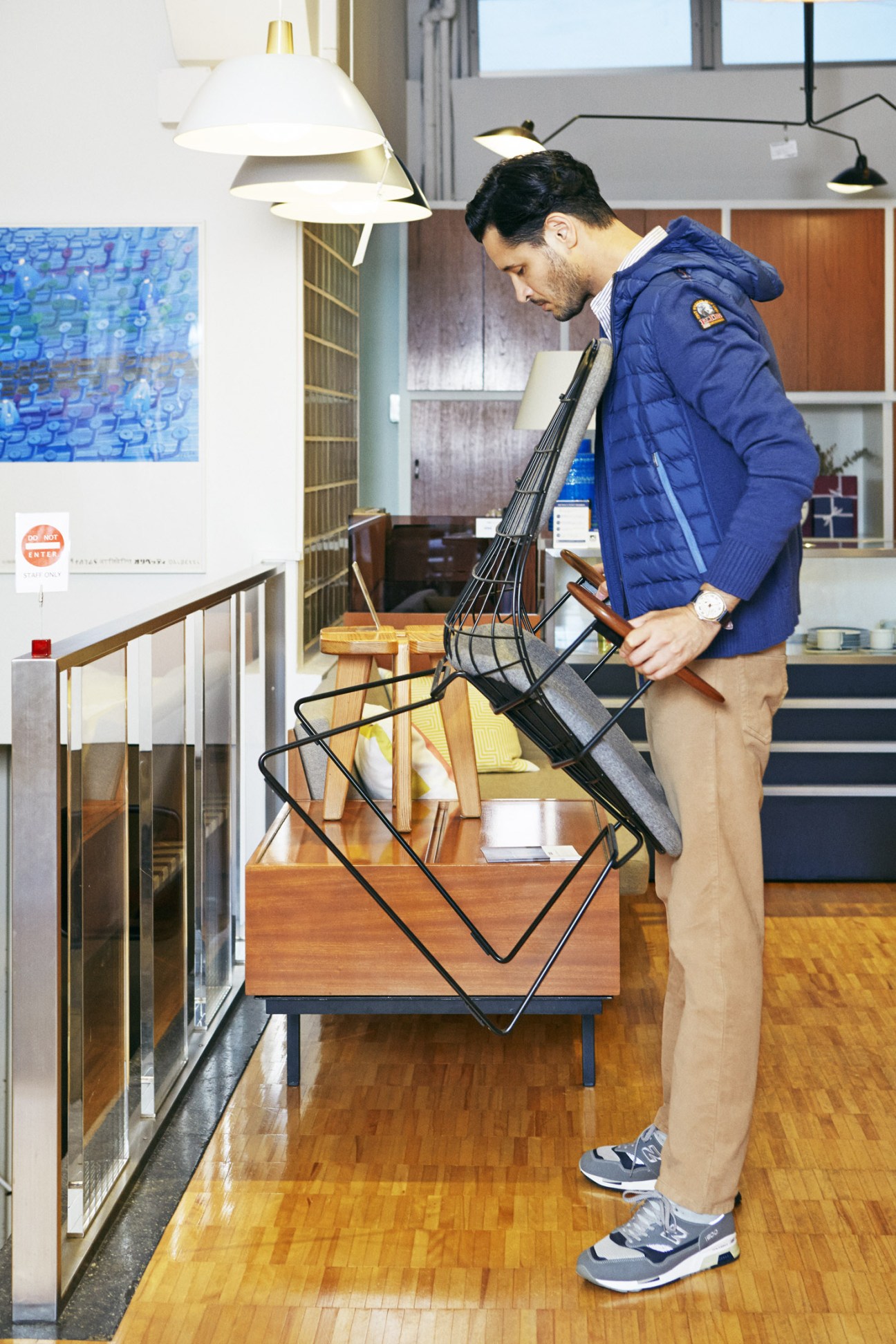 Man in navy Parajumpers jacket and Hackett London trousers arranging modern furniture in minimalist interior workspace.