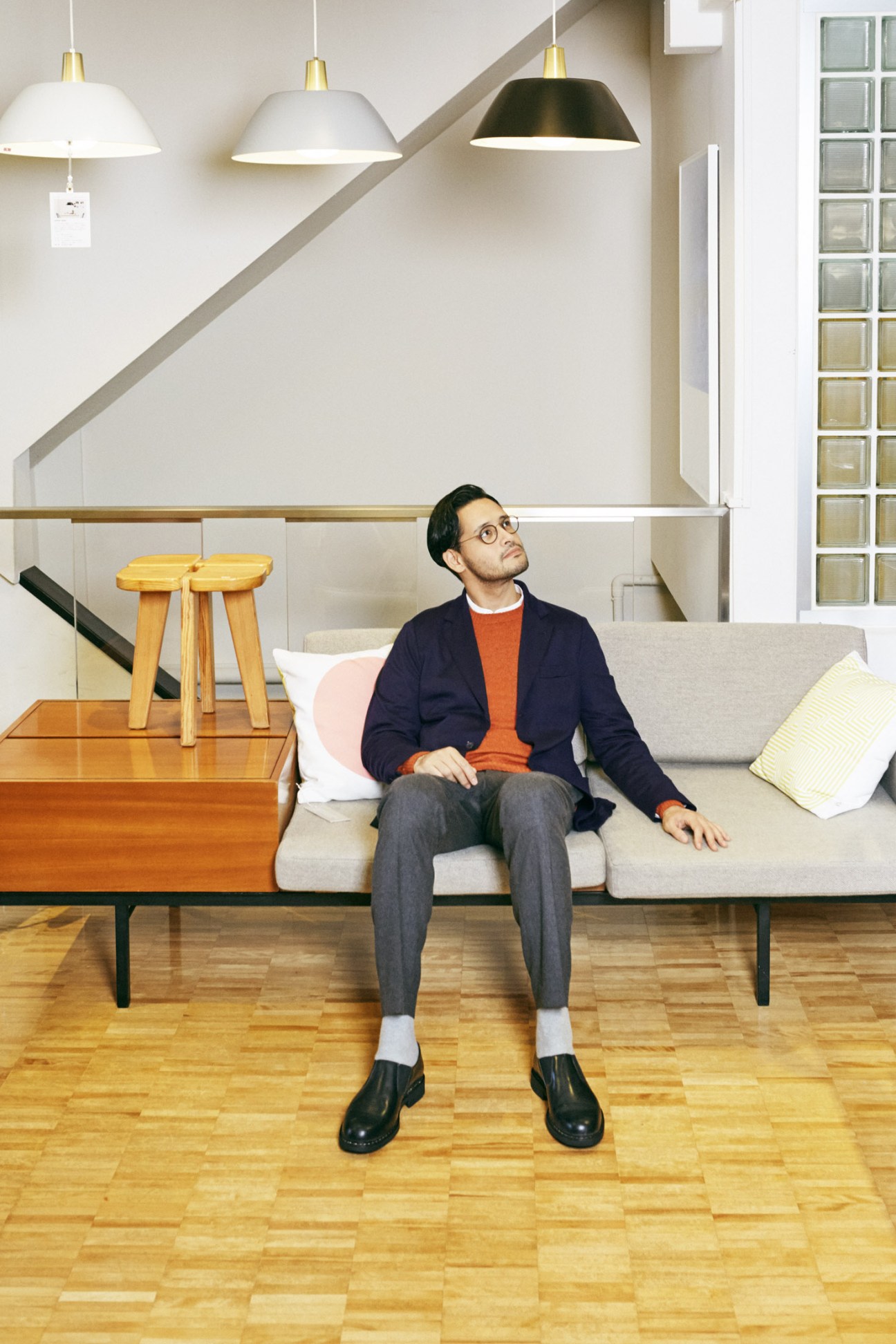 Man wearing navy jacket and orange shirt sitting on modern sofa in minimalist interior with pendant lights.