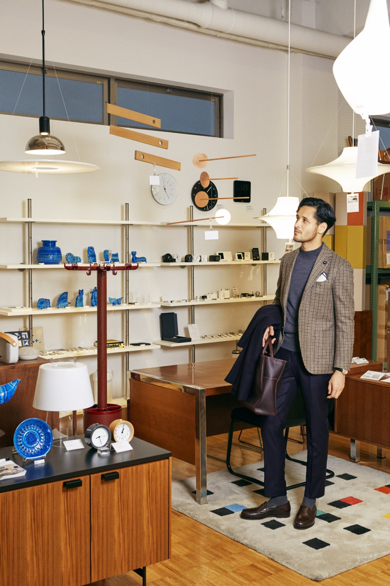 Man in tailored blazer and trousers standing in a modern showroom with shelving and design objects.