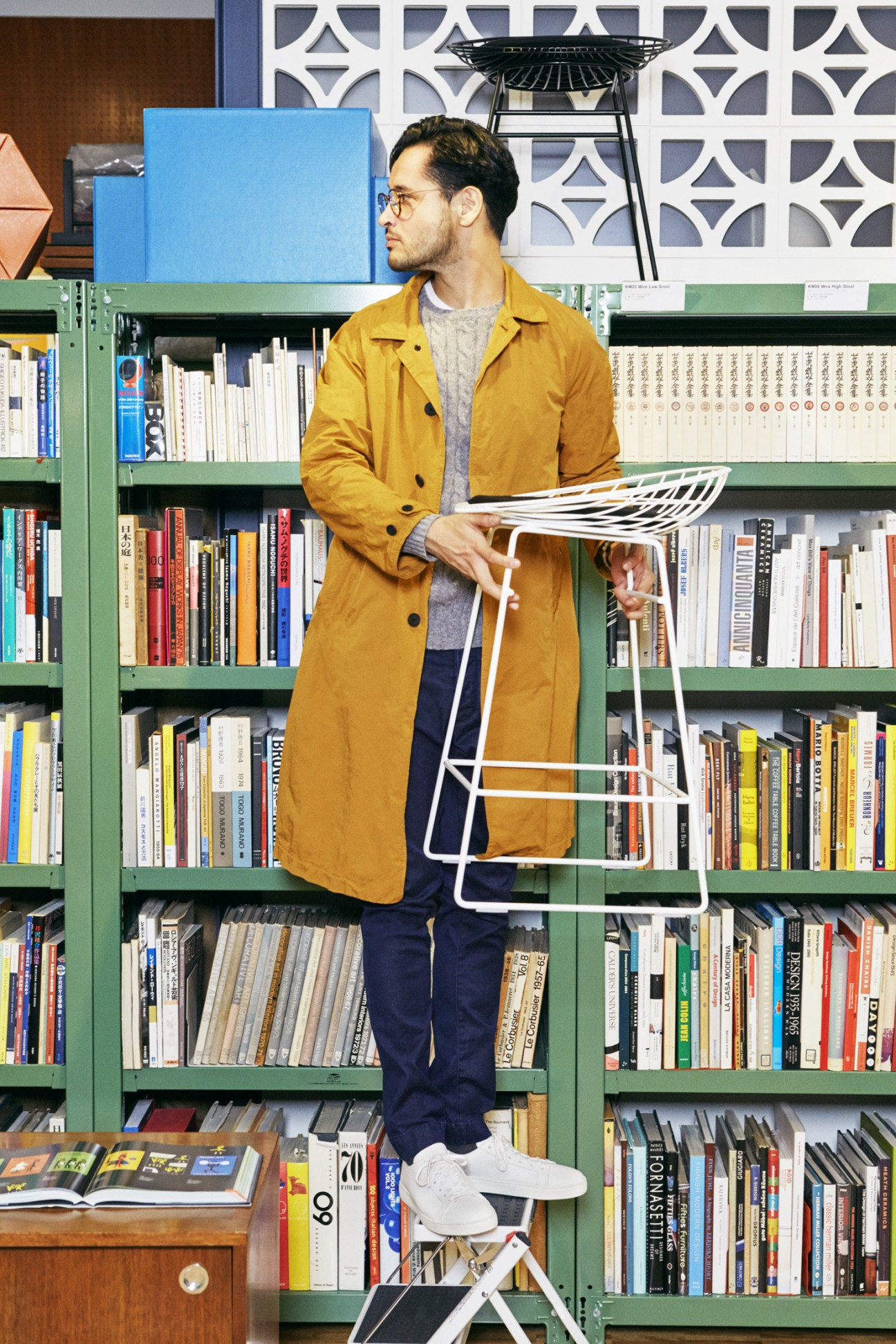 Man in mustard coat and navy jeans standing against green shelving unit holding magazine