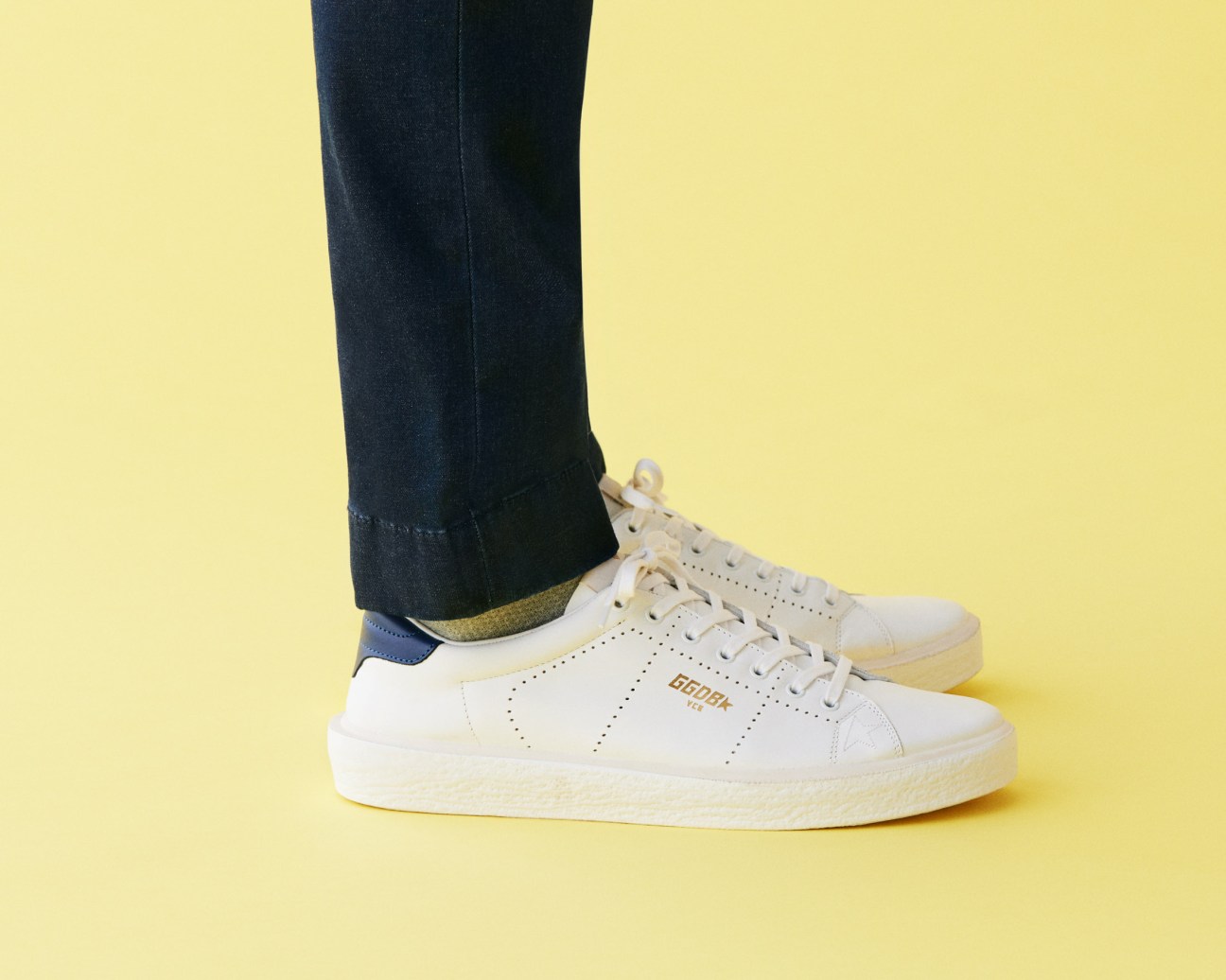 Person wearing dark jeans, white Golden Goose sneakers with navy heel tab, and Uniqlo socks against yellow background.