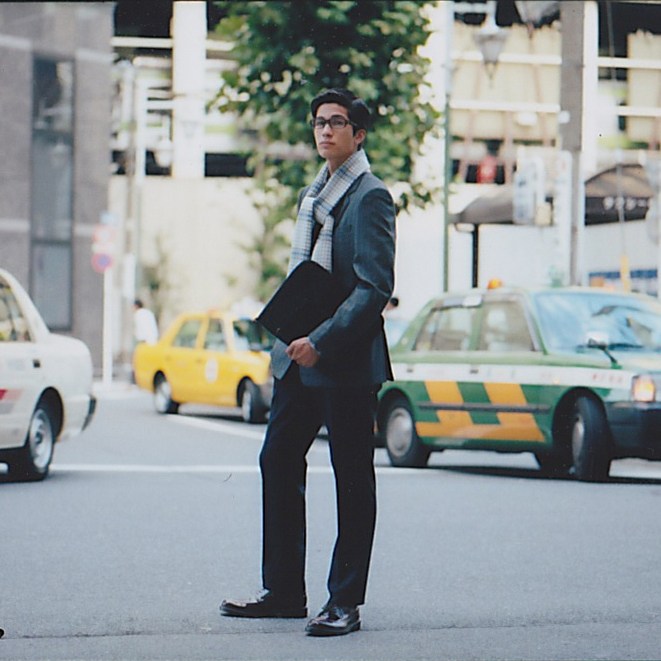 Man in business attire holding a document case stands on a city street with taxis in the background.