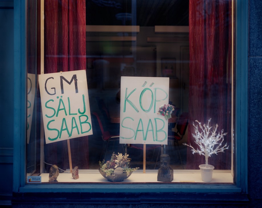 Window display with handmade signs reading "GM SALJ SAAB" and "KÖP SAAB" in green text, surrounded by decorative items.