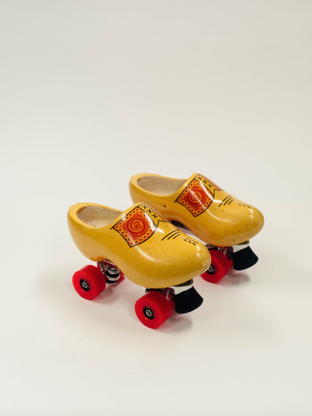 Pair of yellow wooden Dutch clogs with hand-painted red floral decoration mounted on red roller skate wheels.
