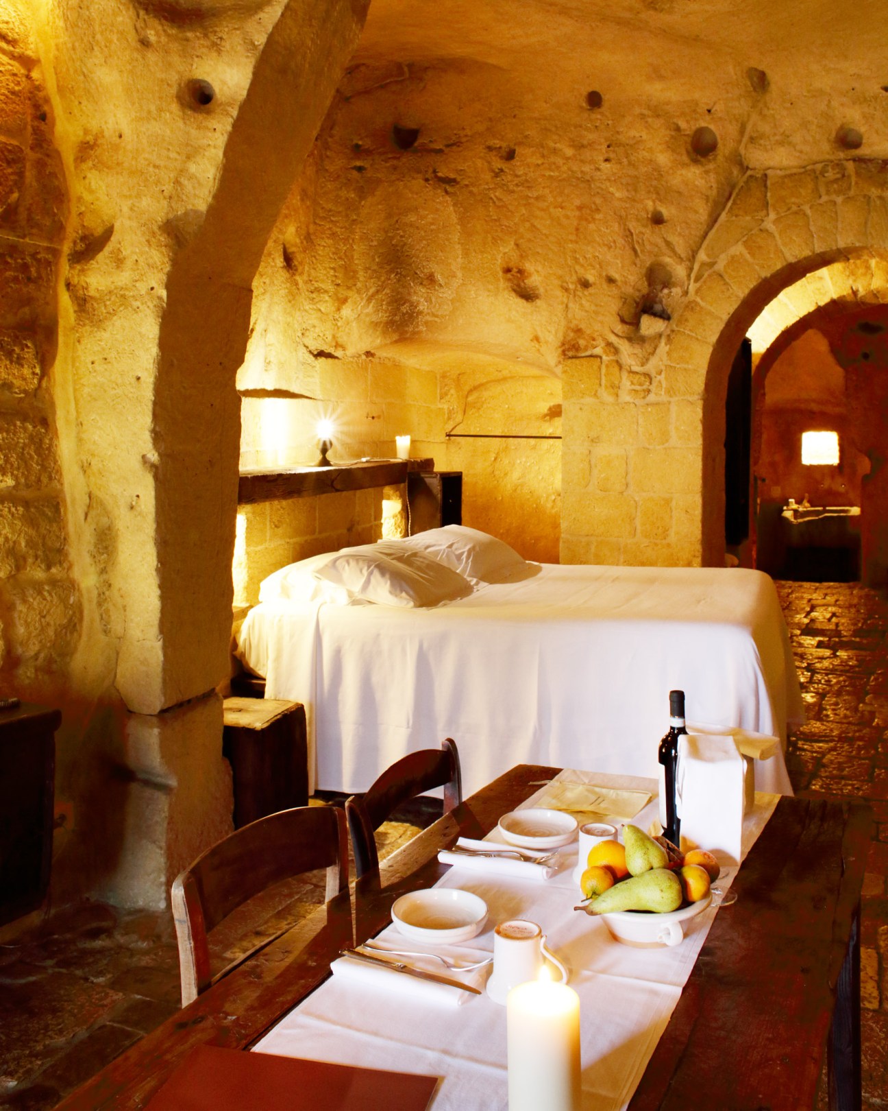 A luxurious cave suite at Sextantio with limestone walls, a white bed, dining table with candles and fruit, warm ambient lighting.