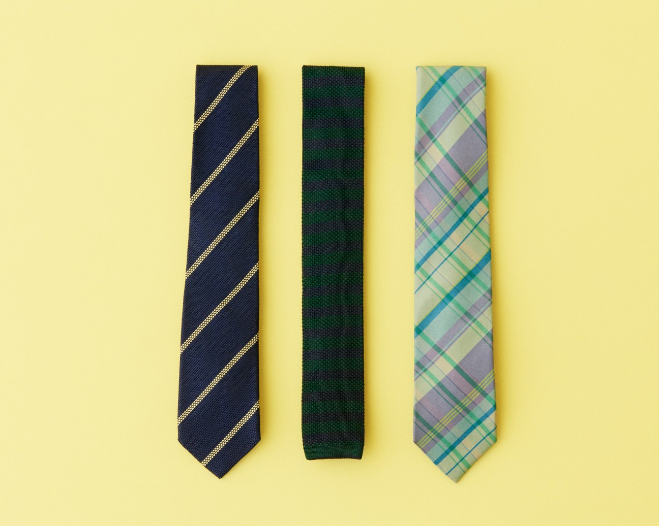 Three neckties displayed on a yellow background: navy with gold stripes, solid green knit, and blue-green plaid.