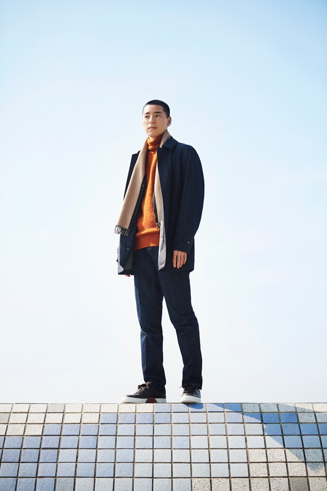 Model wearing layered winter menswear in neutral tones including coat, rollneck jumper and scarf