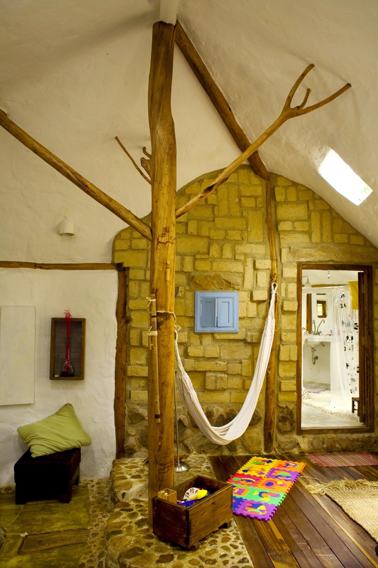 Interior room with white hammock suspended from a large wooden tree branch structure against a yellow stone wall.