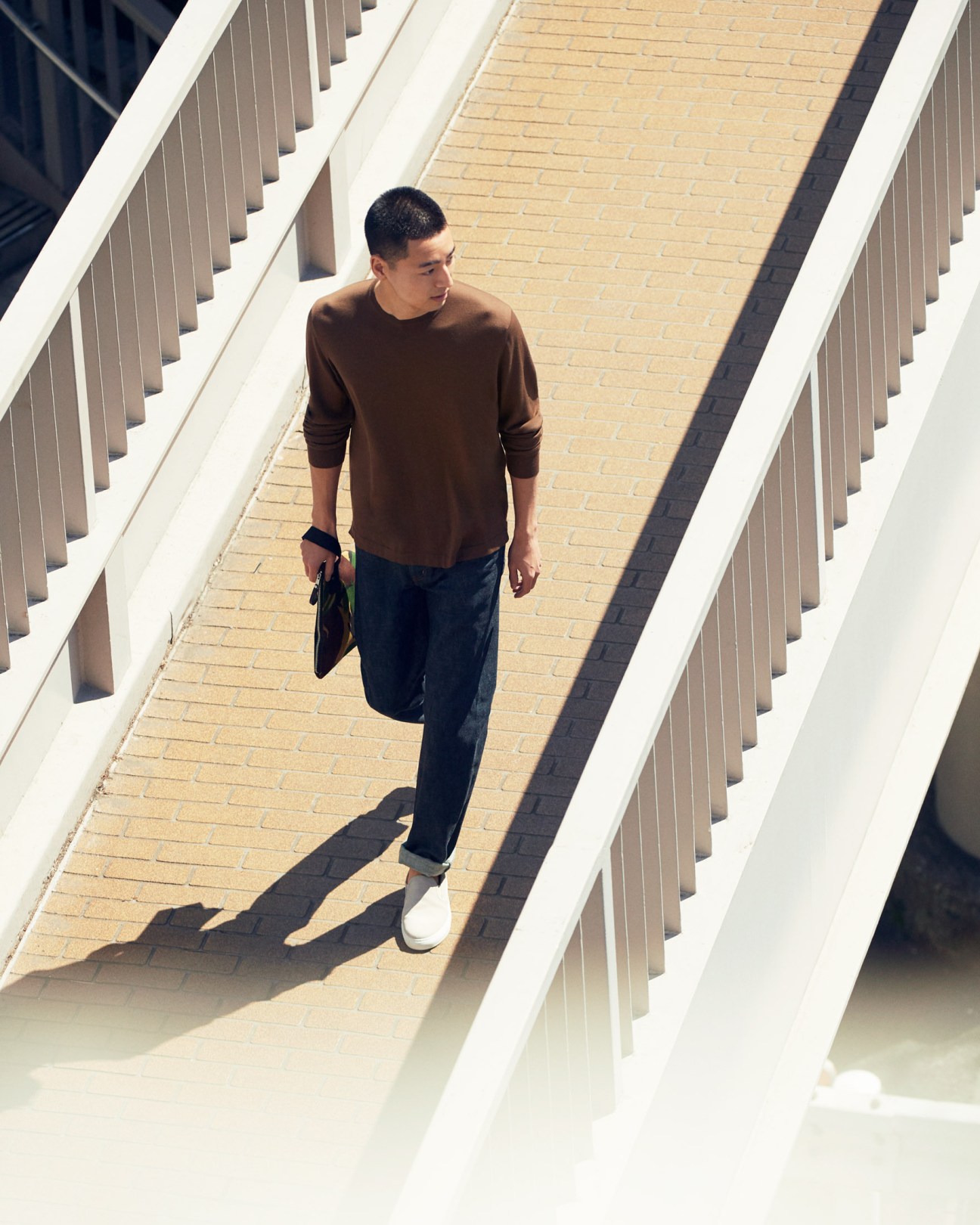 Man wearing brown jumper and dark trousers walking on pedestrian bridge with white railings.