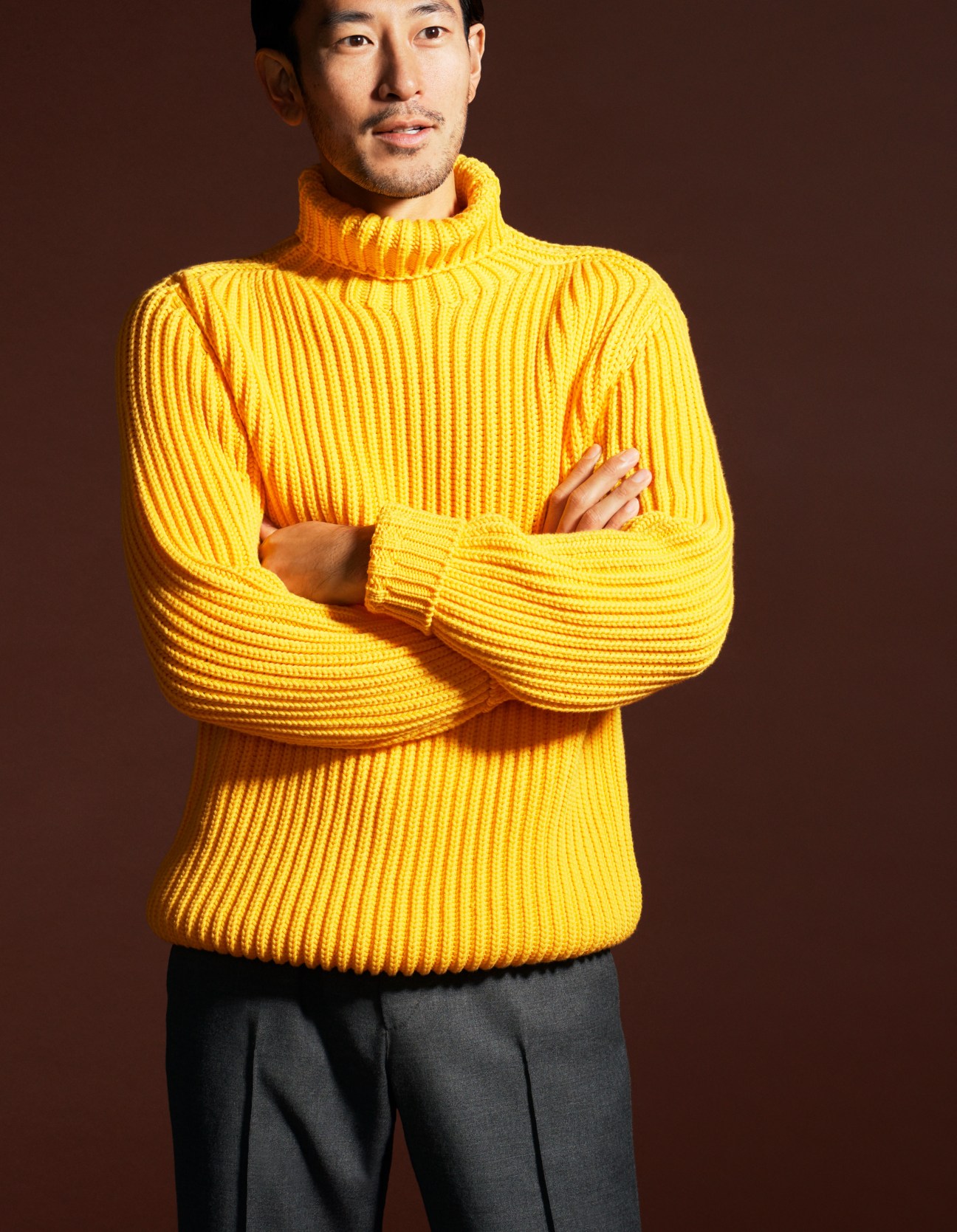 Man in bright yellow ribbed turtleneck sweater with dark trousers standing with crossed arms.