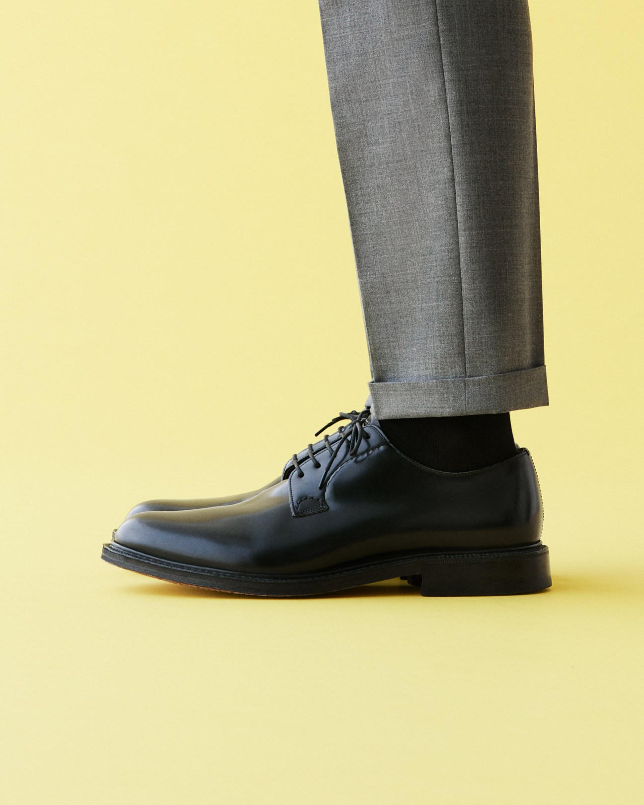 Man's lower legs wearing grey trousers, black socks, and black leather Oxford shoes against yellow background.