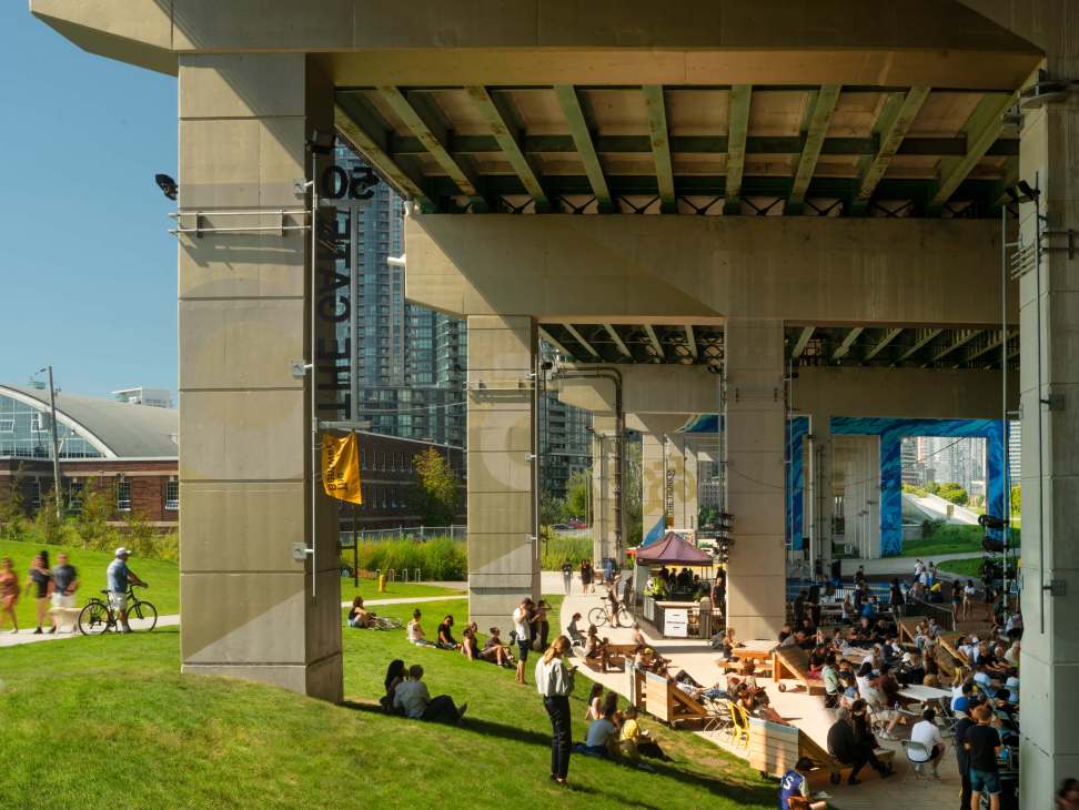 The Bentway in Toronto, a public space beneath the Gardiner Expressway with green lawns, concrete infrastructure, and crowds of people gathering.