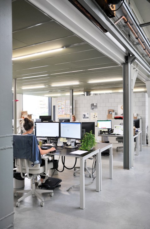 Open office workspace with white desks, computer monitors, office chairs, and exposed ceiling structure