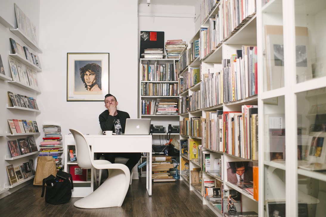 Josh Carey sits at a white desk in Bondi Books, surrounded by white shelves filled with rare books and a framed portrait on the wall.