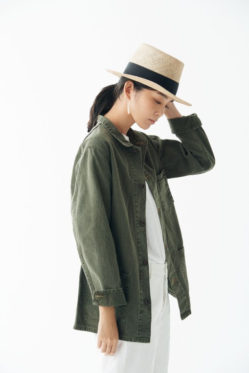 Woman wearing olive denim jacket, white t-shirt, white trousers, straw hat with black band, and dangling earrings.