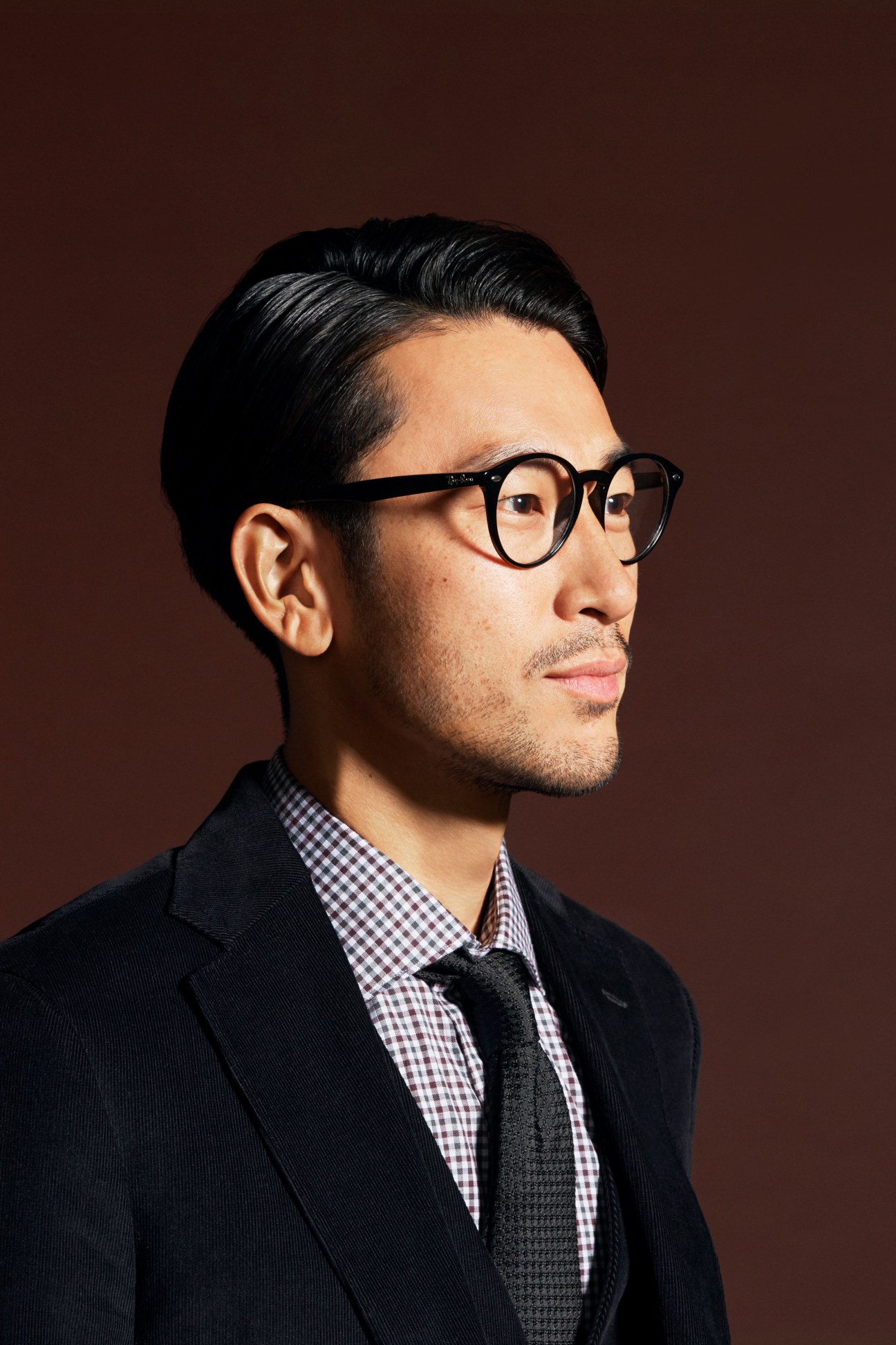Man in grey blazer with white shirt and striped tie wearing Ray-Ban glasses against brown background