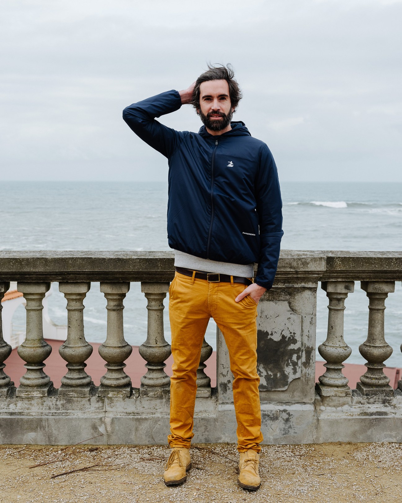 Man wearing navy Parlementia windcheater and mustard trousers standing by ornate balustrade overlooking the sea.