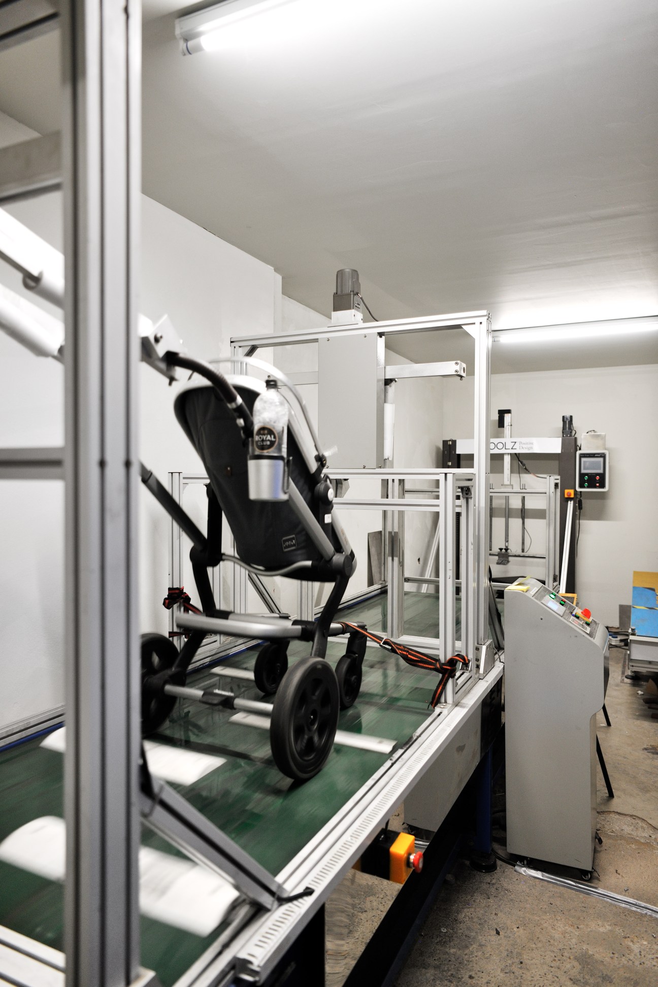 Black pushchair being tested on a green conveyor belt in a white laboratory testing room