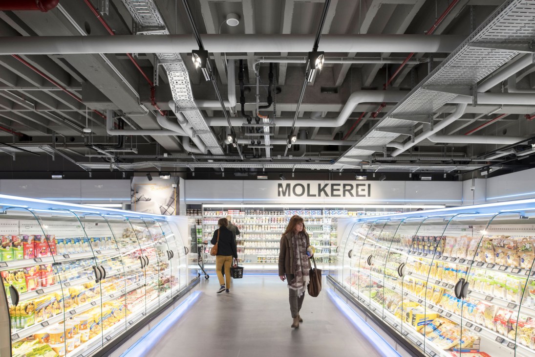Dairy aisle at Zurheide supermarket with illuminated refrigerated display cases on both sides and customers shopping.