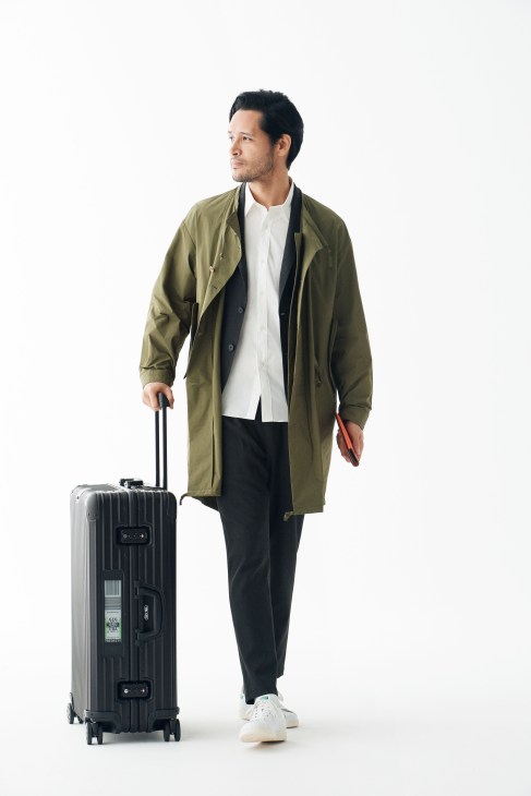 Man in olive coat and black trousers holding Rimowa suitcase against white background.