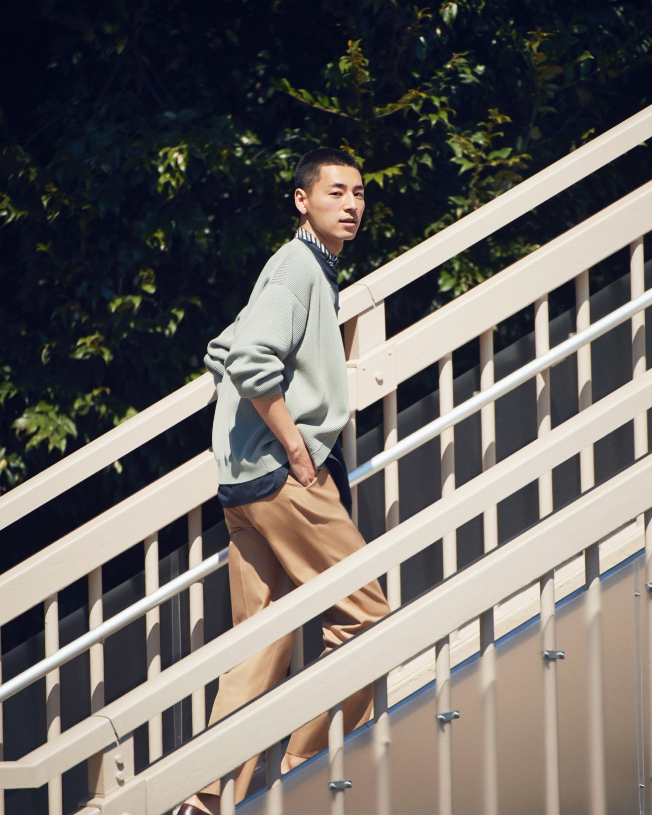 Man wearing light cardigan, trousers, and shoes on a white pedestrian bridge with greenery behind.