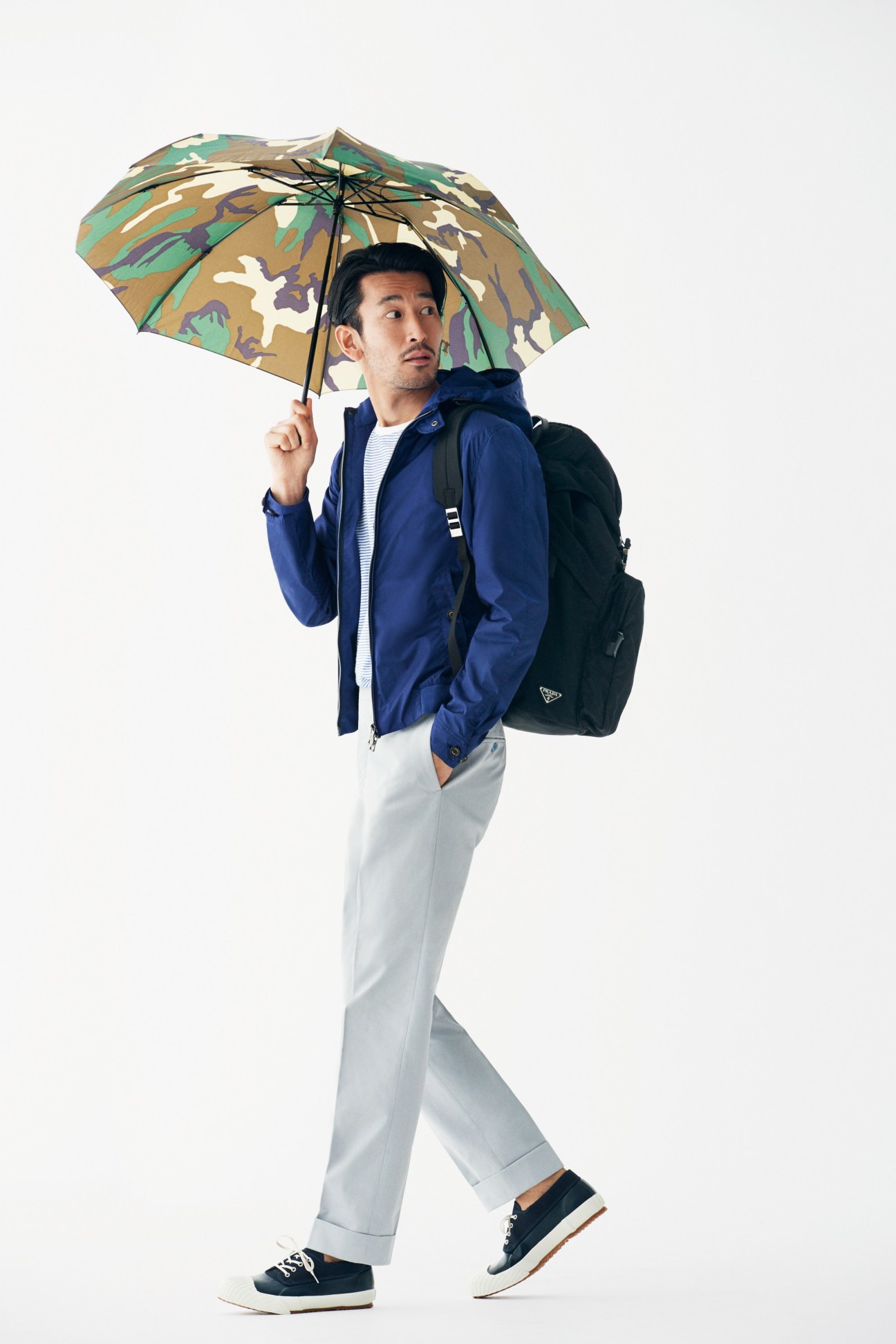 Man holding patterned umbrella wearing navy Herno coat, white pants, and Eytys trainers with backpack.