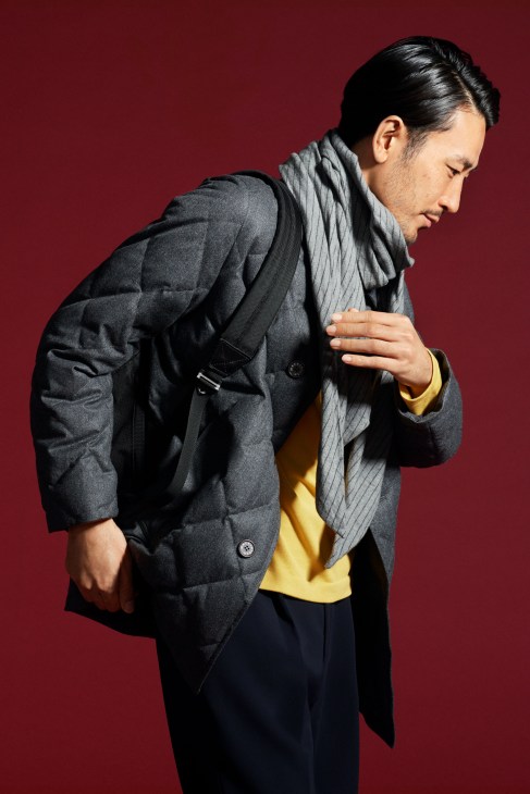 Man in khaki trench coat with grey sweater and scarf carrying black backpack standing against white background