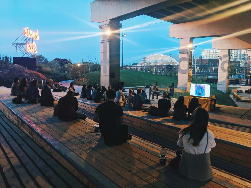 People gather on wooden decking beneath a concrete expressway overpass at dusk, with a neon sign and green space visible in the distance.
