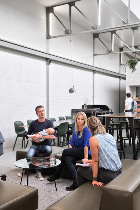 Man holding baby sits across from two women in modern office with industrial ceiling and green chairs