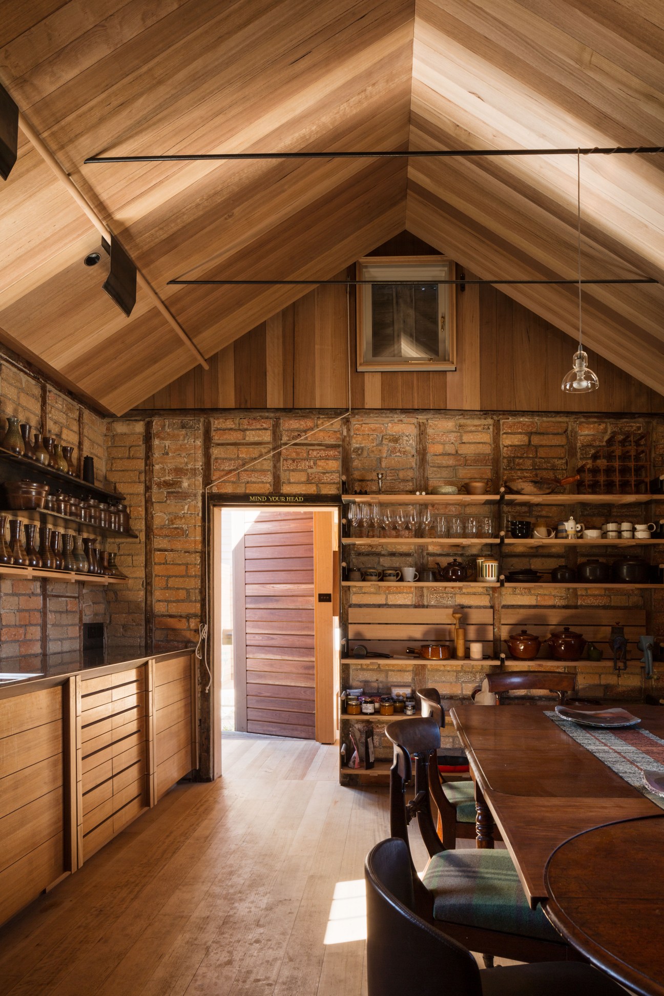 Historic kitchen with gabled wooden ceiling, exposed brick walls, oak cabinetry, and open shelving displaying ceramics.