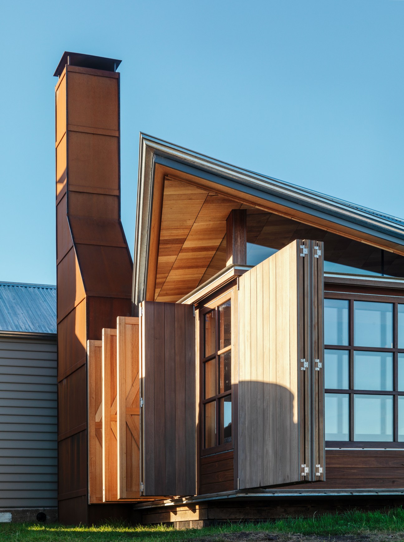 Modern wooden cottage with retractable shutters and tall brown metal chimney against clear blue sky.