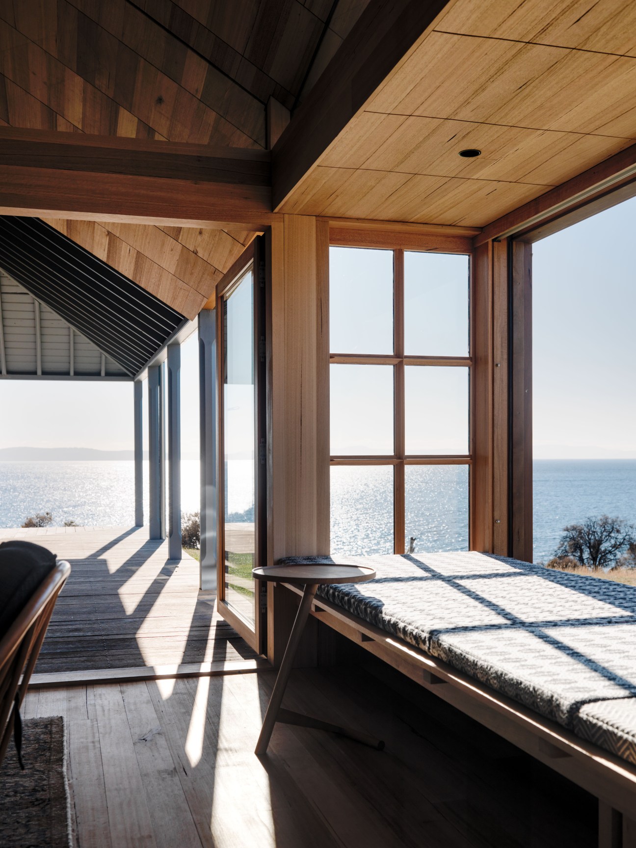 Interior of a wood-lined room with large windows overlooking the ocean, featuring a daybed window seat with cushioning and modern wooden beams.