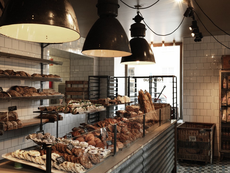 Interior of Fabrique bakery in Stockholm with white subway tiles, black industrial pendant lights, shelves of brown baked goods, and a counter display of pastries and sourdough loaves.