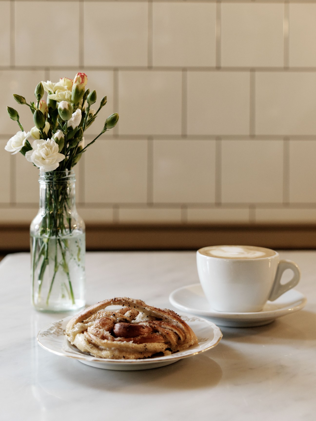White cinnamon pastry and cappuccino on a table with white subway tile wall and fresh carnations in a glass vase.