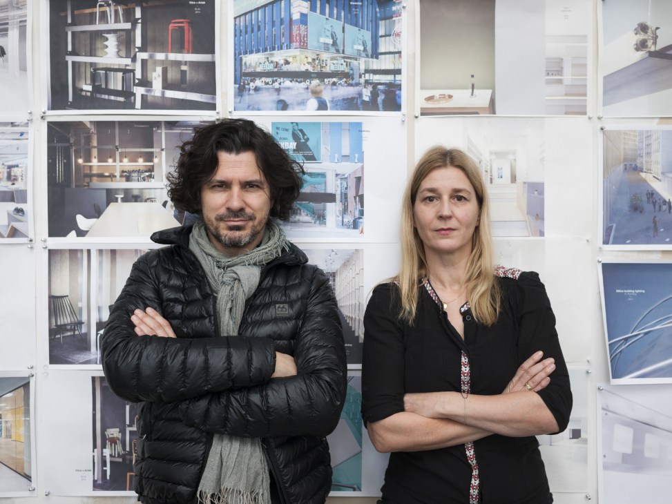 Two architects stand with arms crossed in front of architectural drawings and renderings pinned to a white wall.