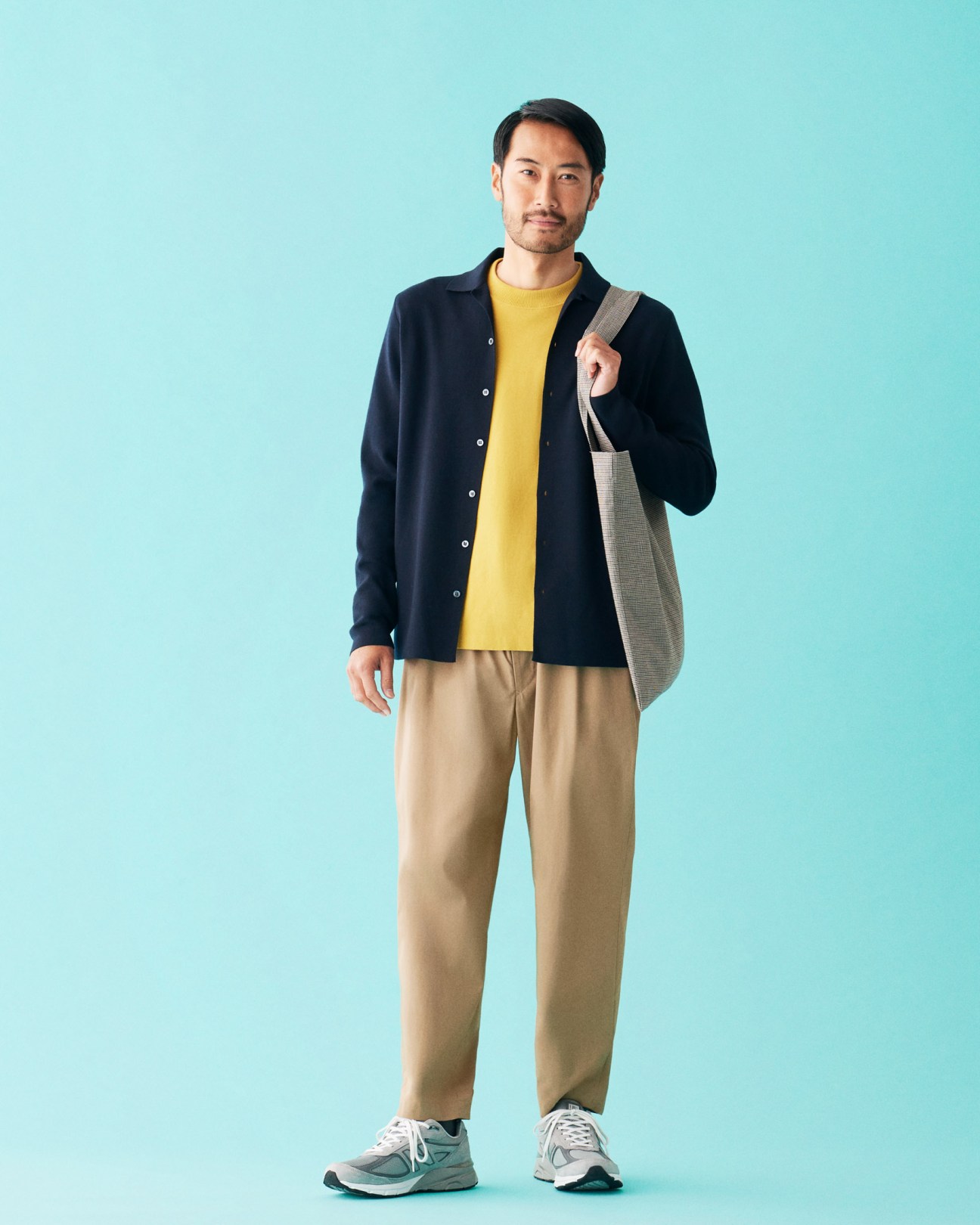 Man wearing navy cardigan, yellow jumper, beige trousers, and New Balance trainers against light blue background.