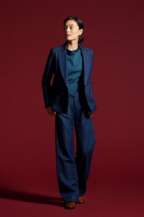 Woman in brown blazer and matching trousers with white shirt and black shoes wearing earrings standing against white background