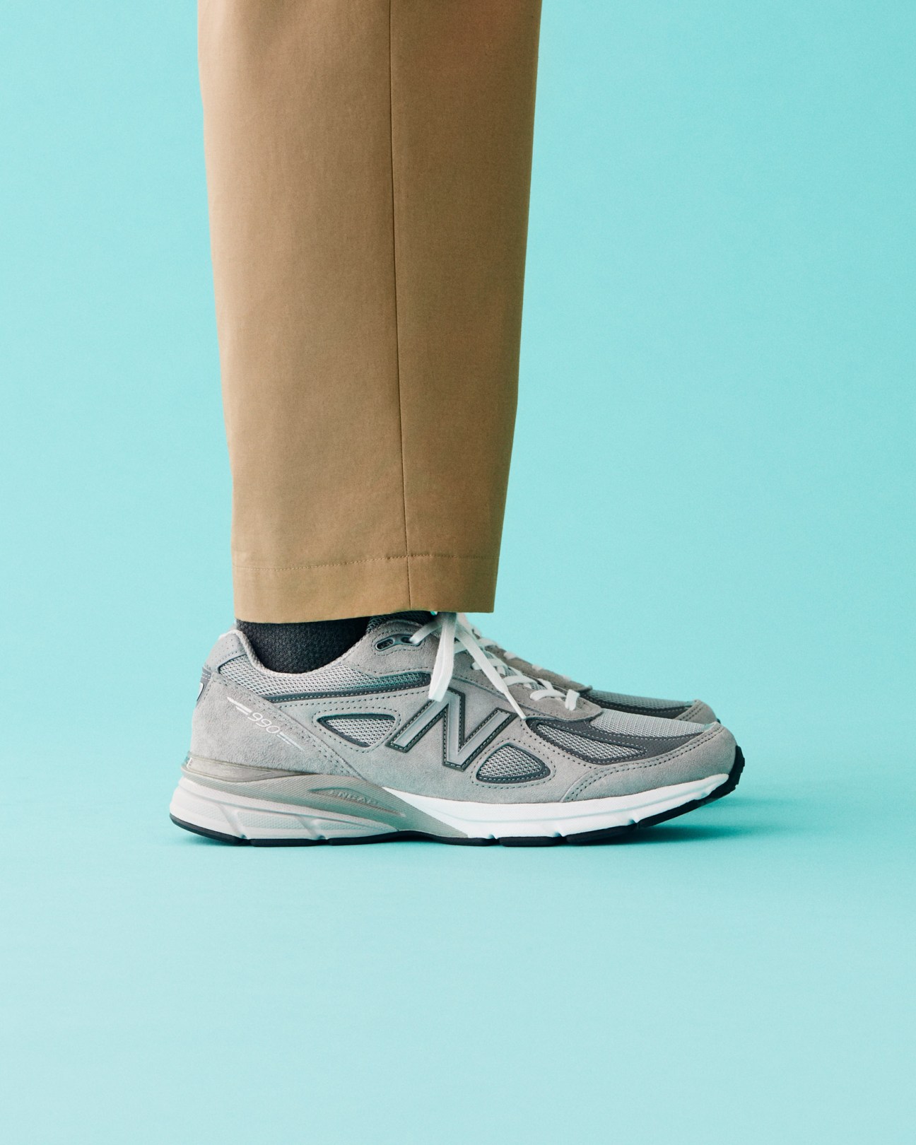 Person wearing beige trousers, dark socks, and grey New Balance trainers against a turquoise background.