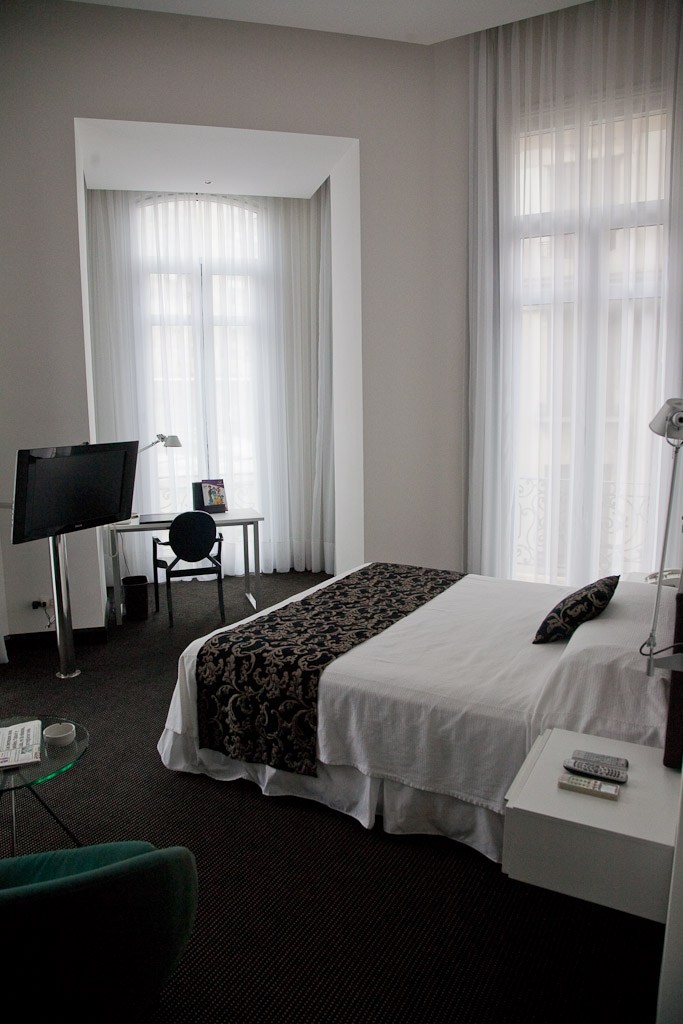 Modern hotel room at Plaza Fuerte Hotel with white walls, bed with black patterned runner, desk with TV, and floor-to-ceiling sheer white curtains.